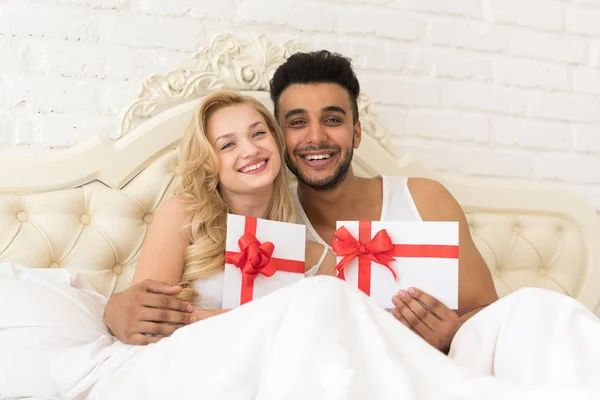 Joven pareja feliz acostada en la cama, el hombre y la mujer hispanos sostienen sobre presente con cinta, celebración del aniversario — Foto de Stock