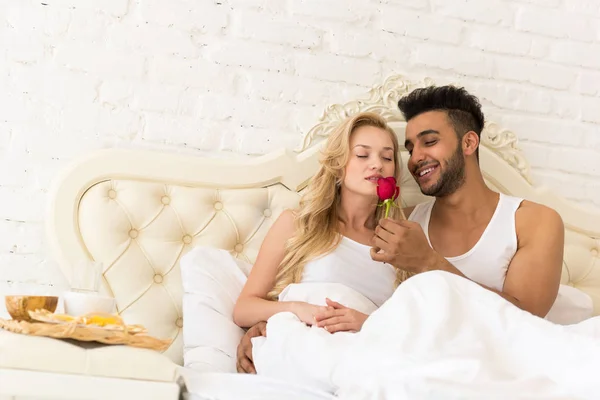 Pareja joven acostada en la cama desayunar por la mañana con flor de rosa roja, feliz sonrisa hispana hombre y mujer —  Fotos de Stock