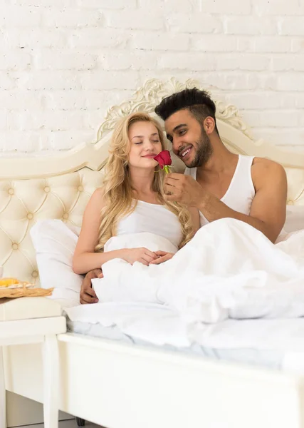 Mladý pár leží v posteli jíst snídani ráno s červený květ růže, šťastný úsměv hispánský muž a žena — Stock fotografie