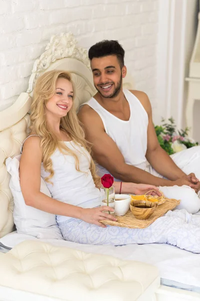 Junges Paar im Bett liegend frühstückt morgens mit roter Rosenblüte, glückliches Lächeln spanischer Mann und Frau — Stockfoto