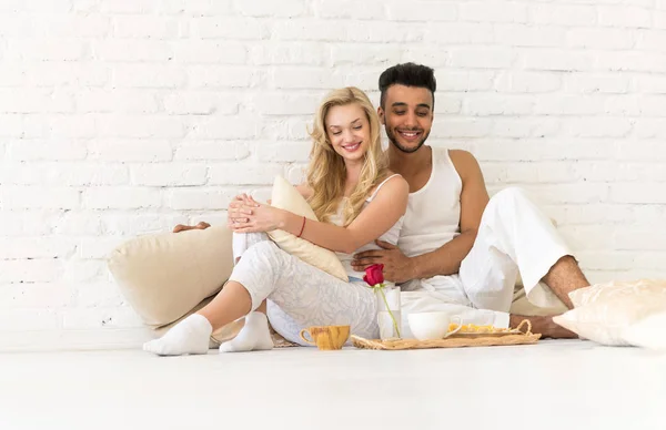 Pareja joven se sienta en el piso de almohadas, feliz hombre hispano y mujer bandeja de desayuno amantes en el dormitorio —  Fotos de Stock