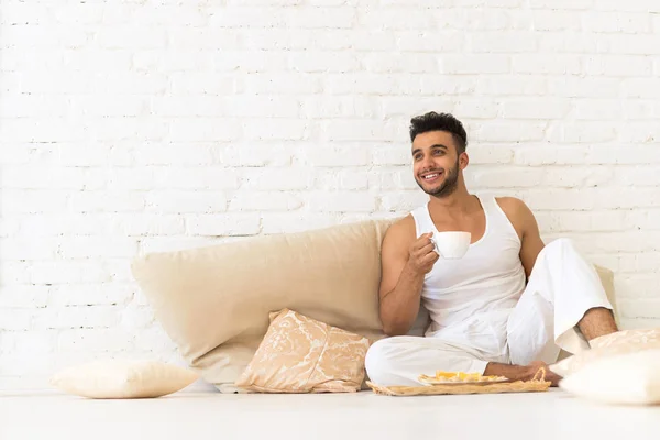 Jonge Spaanse Man zit op vloer kussens, gelukkig Glimlachende man met ontbijt lade Look kopie ruimte slaapkamer — Stockfoto