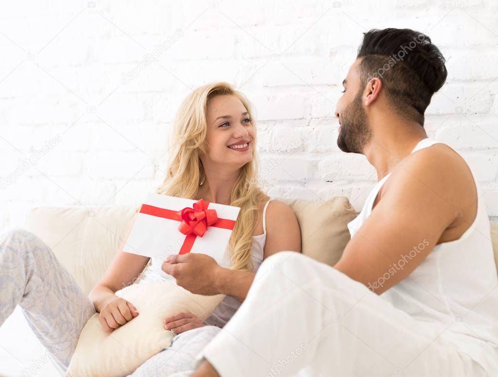 Young Couple Sit On Pillows Floor, Happy Smile Hispanic Man Woman Hold Present Envelope With Ribbon Lovers In Bedroom