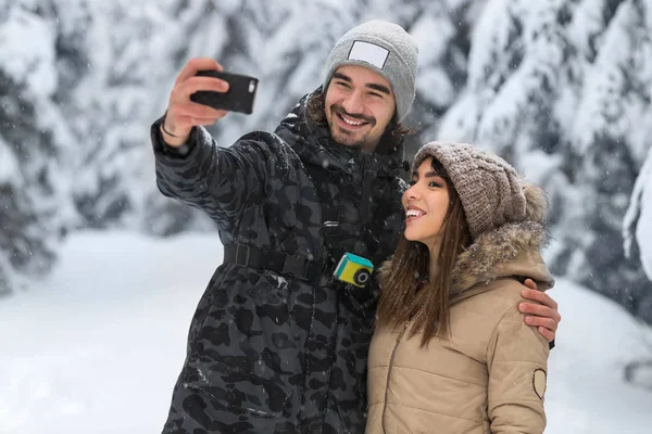 Az ember tartsa intelligens telefon kamera véve Selfie fénykép fiatal romantikus pár mosoly hó erdő szabadtéri — Stock Fotó