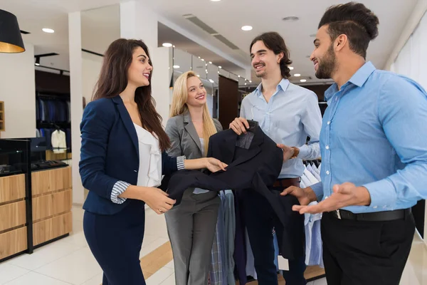 Due assistenti di negozio femminili che offrono ai giovani uomini d'affari una nuova tuta per lo shopping nel negozio di abbigliamento maschile di lusso — Foto Stock