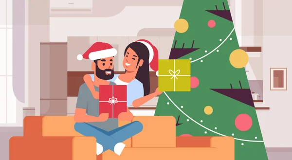 Couple holding gift boxes merry christmas happy new year holiday celebration concept man woman embracing wearing santa hats sitting on couch near fit tree modern living room interior horizontal — Stock Vector