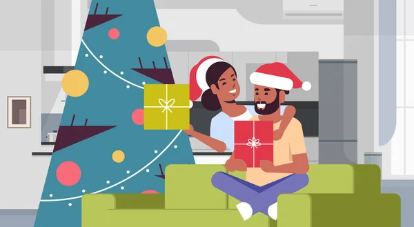 Pareja celebración de cajas de regalo feliz navidad feliz año nuevo celebración concepto hombre mujer abrazando el uso de sombreros de santa sentado en el sofá cerca del árbol en forma moderna sala de estar interior horizontal — Vector de stock