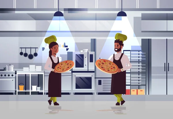 专业厨师夫妇拿着托盘与新鲜的比萨饼非洲美国男子女人在制服站在一起烹饪食品概念现代餐厅厨房内部水平全长 — 图库矢量图片