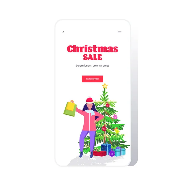Flicka i Santa hatt innehav shoppingväskor stående nära gran jul försäljning helgdagar firande koncept smartphone skärm online mobil app full längd gratulationskort — Stock vektor
