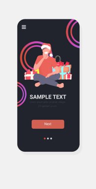 Noel Baba şapkalı adam hediye kutularının yanında oturuyor. Mutlu Noeller, yeni yıl kutlamaları, akıllı telefon ekranı, mobil uygulama, dikey çizim.