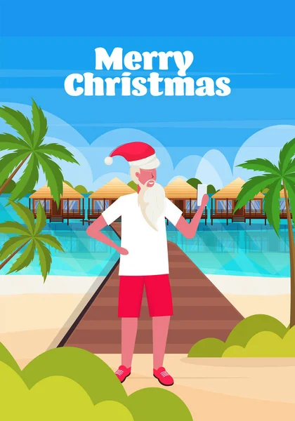Mężczyzna sobie Santa Claus kapelusz za pomocą smartfona na tropikalnej plaży Nowy rok święta wakacje koncepcja krajobraz morski tło kartka okolicznościowa pełna długość pionowa — Wektor stockowy