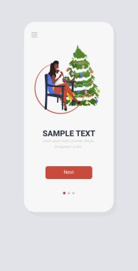 Afrikalı Amerikalı bir kadın Noel ağacının yanındaki koltukta oturuyor. Yılbaşı bayramı kutlaması konsepti. Akıllı telefon ekranı mobil uygulaması.