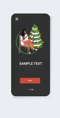 Afrikalı Amerikalı bir kadın Noel ağacının yanındaki koltukta oturuyor. Yılbaşı bayramı kutlaması konsepti. Akıllı telefon ekranı mobil uygulaması.