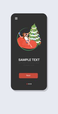 Cep telefonlu bir kadın noel ağacı yakınında noel kutlaması konsepti akıllı telefon ekranı mobil uygulaması tam boy dikey