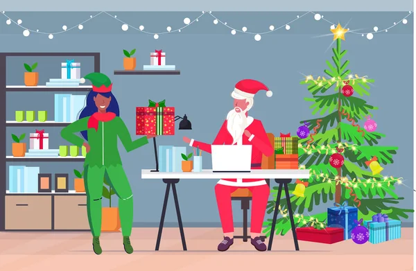 Санта Клаус використовує ноутбук афроамериканська жінка-ельф помічник тримає подарункову коробку різдвяні новорічні свята концепція святкування сучасний офіс внутрішній вітальна листівка горизонтальна повна довжина — стоковий вектор
