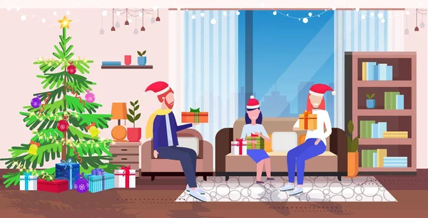 Lycklig familj i Santa sitter på soffa nära gran jul nyår semester firande koncept föräldrar och dotter håller presentförpackning vardagsrum interiör full längd horisontellt — Stock vektor
