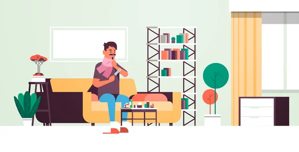 Sick man measuring temperature with thermometer unhealthy guy in scarf suffering from cold flu virus sitting on sofa illness concept modern living room interior flat full length horizontal — Stock vektor