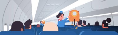 Hostes yolculara acil durumlarda ceket can yeleklerinin nasıl kullanılacağını anlatıyor. Kadın uçuş görevlileri güvenlik gösterimi konsepti modern uçak panosu iç yatay portresi.
