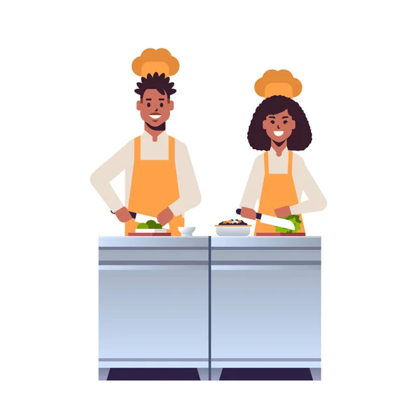 Cocina pareja de chefs profesionales picando verduras frescas en la tabla de tallar afroamericano hombre mujer restaurante trabajadores en uniforme preparando ensalada cocina concepto de comida plana de longitud completa — Vector de stock