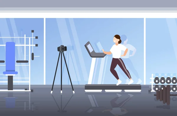 Žena blogger běží na běžícím pásu nahrávání videa s kamerou na stativ sociální sítě blogování zdravý životní styl koncept moderní tělocvična interiér plná délka horizontální — Stockový vektor