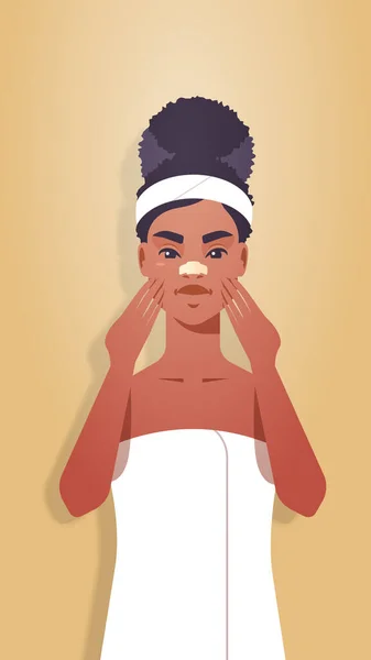 Mujer joven aplicando crema para la piel vestida con toalla chica afroamericana limpiando y cuidando su rostro tratamiento facial spa tratamiento facial retrato vertical — Archivo Imágenes Vectoriales