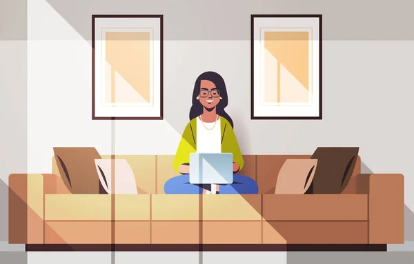 Vacker indian kvinna sitter på soffan med laptop flicka som arbetar hemifrån frilans koncept modernt vardagsrum interiör horisontellt full längd — Stock vektor