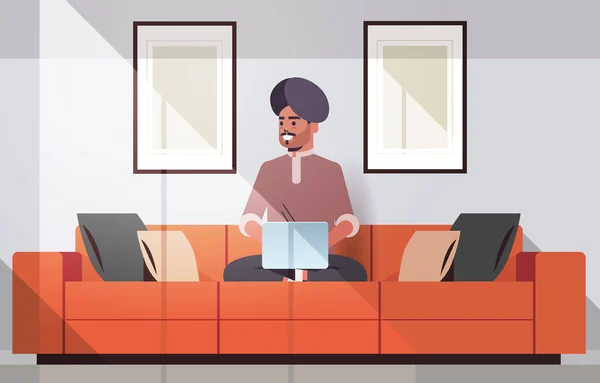 头戴头巾的印地安人坐在沙发上使用笔记本电脑，他是根据家庭自由职业者的概念工作的。现代客厅内部横向全长 — 图库矢量图片
