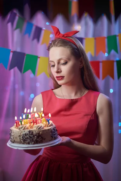 Красивая молодая винтажная девушка в стиле булавки, стоящая в красочной освещенной сцене, держа торт со свечами . — стоковое фото