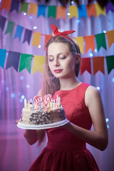 Beautiful young vintage pin up style girl standing in colorful lighted scene holding cake with candles.