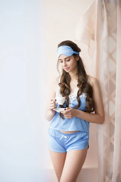 Krásná žena stála u okna s kávou v modrém pyžamovém stroji a šortky s krajkou — Stock fotografie