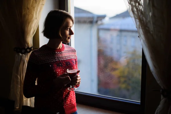 美しい女性 ウィンドウの横に立って 赤冬の服を着て 飲むホットワイン — ストック写真
