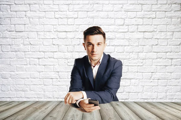 Attractive businessman in trendy suit sitting at desk against empty background — Stock Photo, Image