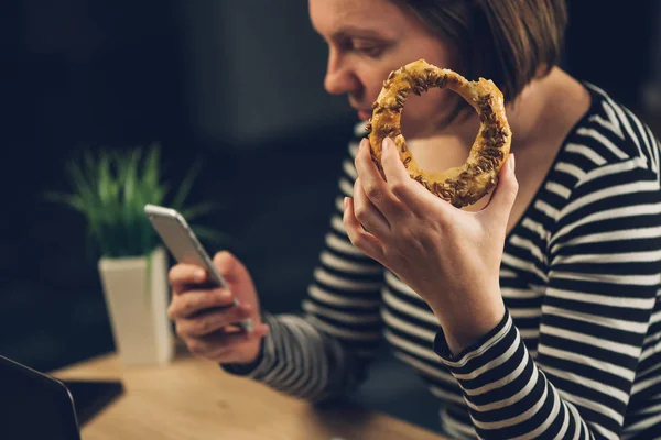Женщина ест бублик с кунжутом и пользуется мобильным телефоном — стоковое фото