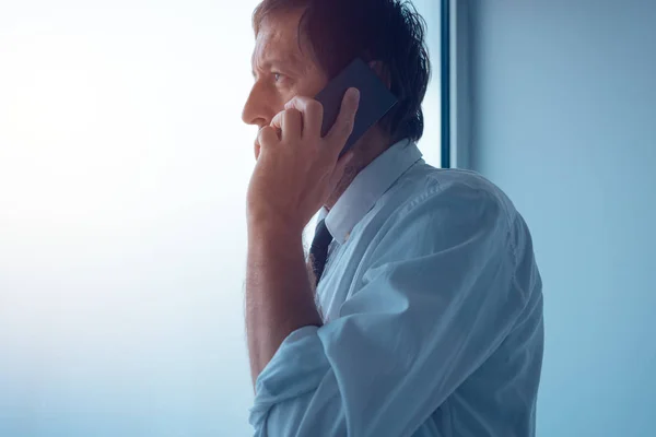 Бизнесмен, стоящий у окна офиса и разговаривающий по мобильному телефону — стоковое фото