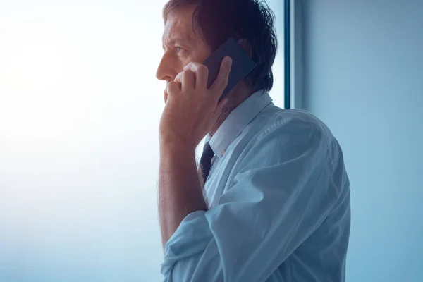 Businessman standing next to office window and talking on mobile — Stock Photo, Image