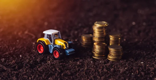 Agricultural tractor toy and golden coins on fertile soil land — Stock Photo, Image