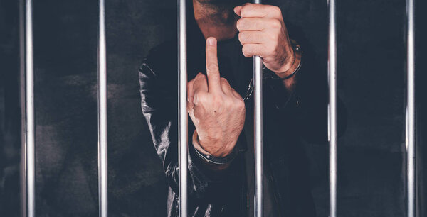 Handcuffed man behind prison bars giving middle finger