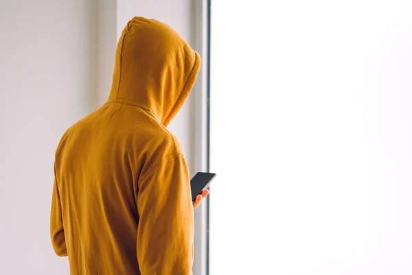 Uomo con cappuccio e telefono cellulare in piedi vicino alla finestra — Foto Stock