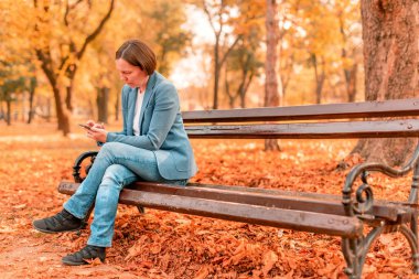 Güz parkındaki bankta cep telefonunda yazı yazan kadın