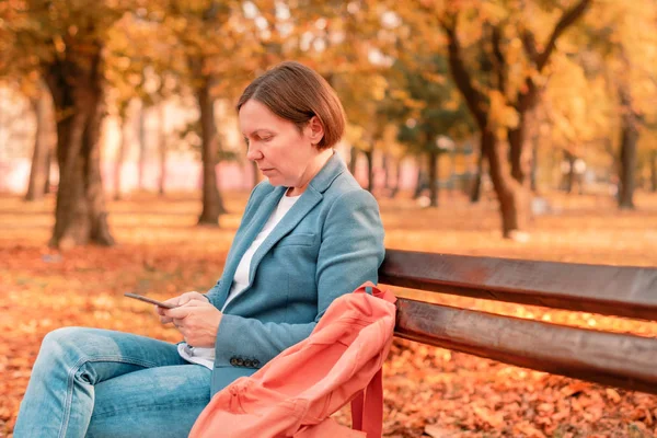 Mulher digitando mensagem de texto no telefone celular no banco do parque — Fotografia de Stock