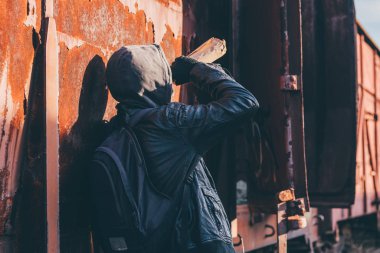 Eski tren vagonunun yanında alkol içen evsiz ayyaş alkolizm konseptinde kapüşonlu sarhoş adam, seçici odaklanma.