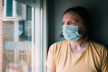 Virüs salgını sırasında kendini izole eden kadın pencereden bakıyor. Koruyucu cerrahi maskeli endişeli kadın, seçici odaklanma