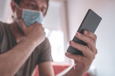 Endişeli adam ev karantinasında cep telefonu kullanıyor. Covid-19 koronavirüs semptomları, seçici odaklanma...