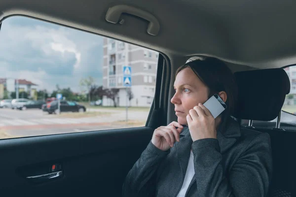 Doubtful businesswoman talking on mobile phone on car back seat, business on the move concept. Elegant adult caucasian female person using smartphone for communication.