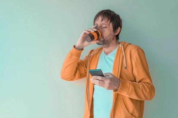 男はコーヒーを飲み 午前中に携帯電話を使用して 黄色のジップパーカーを持つ大人の男性はコーヒーカップとスマートフォンを保持し 選択的な焦点 — ストック写真