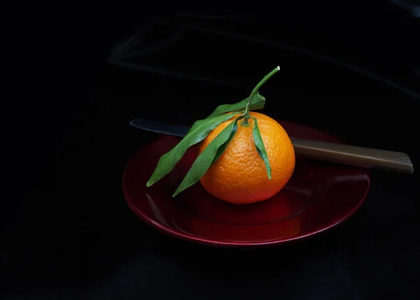 Mandarin type small orange on red plate, with leaves. Whole, unpeeled. With knife, dark background. — 图库照片