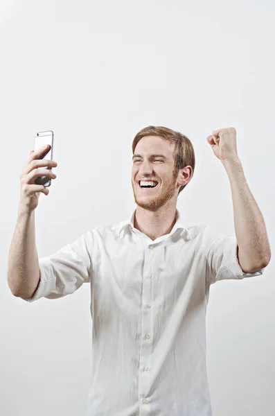 Дуже щаслива, посміхаючись молодий дорослий чоловік у білій сорочці, дивлячись на свій мобільний телефон, Вдивіться, накачуванням кулак — стокове фото