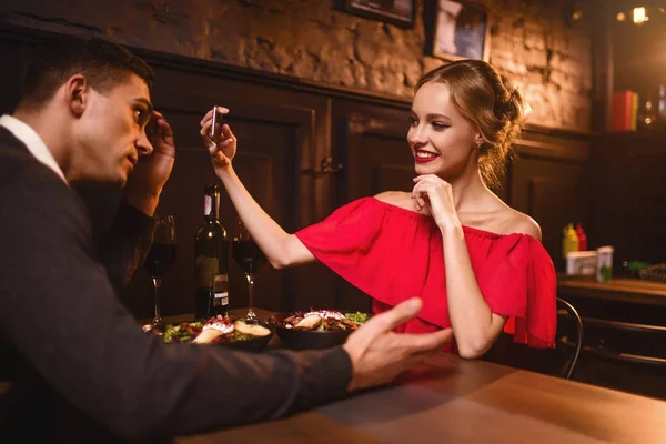 穿着红衣服的微笑的女人在电话摄影机上拍到她男人的照片 年轻的情侣在餐厅 浪漫的约会 典雅的妇女在红色礼服和英俊的人用餐 — 图库照片