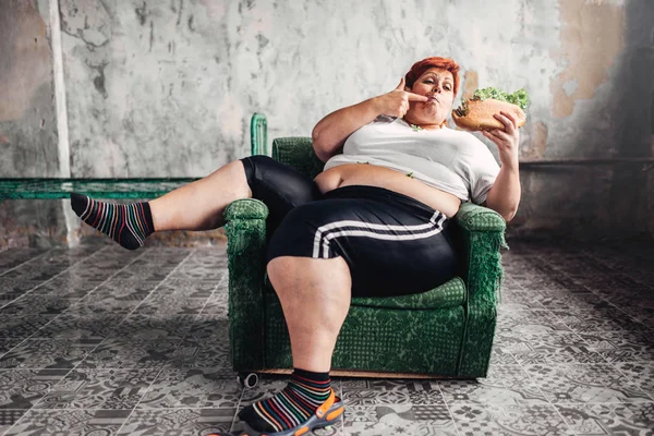fat woman sitting in armchair and eating sandwich, unhealthy lifestyle and obesity concept