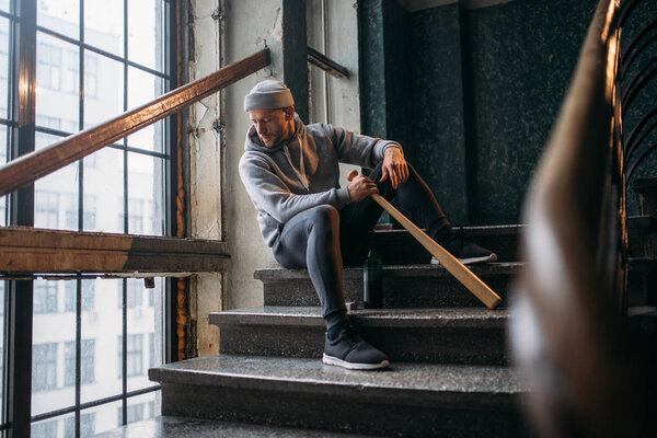 male theif with bottle of alcohol and baseball bat sitting on stairs waiting for victim, crime concept